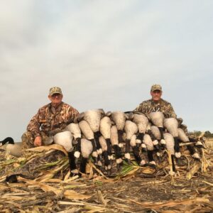 Two men sitting next to a pile of ducks.